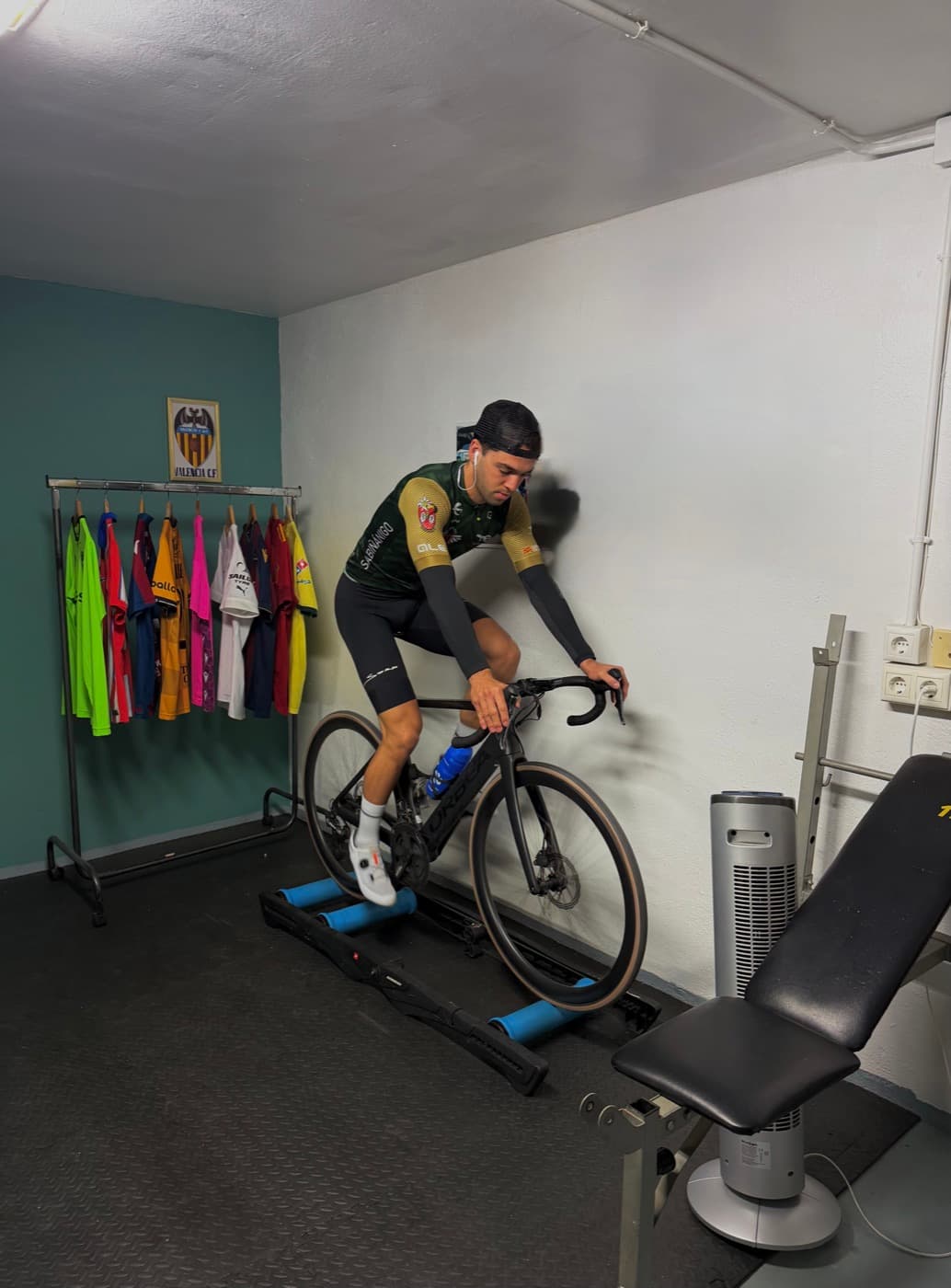 Ciclista entrenando en el rodillo en un garaje, junto a un perchero con maillots